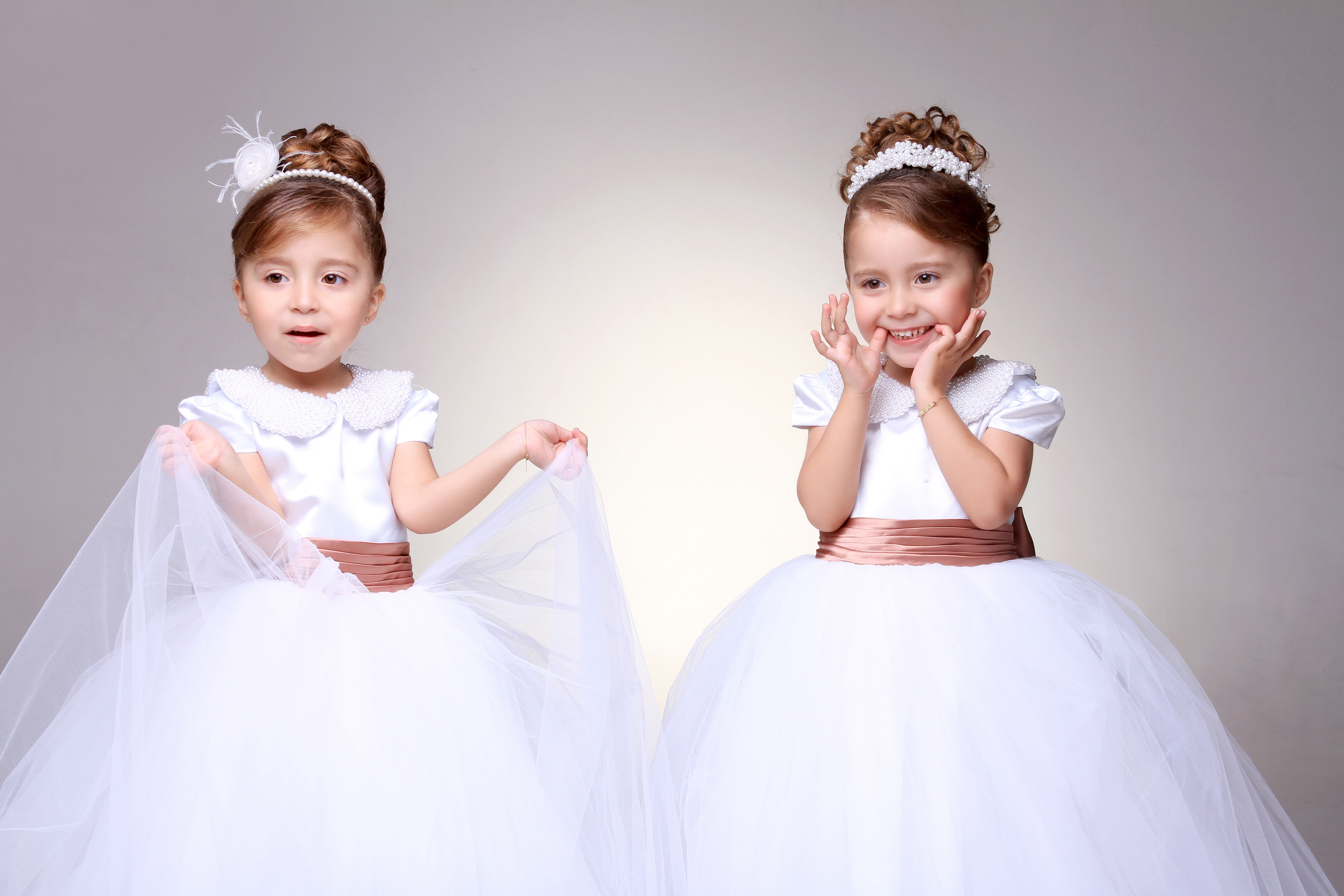 Duas daminhas com penteados de coque cacheado, franja lateral e uma tiara fina de pérolas.