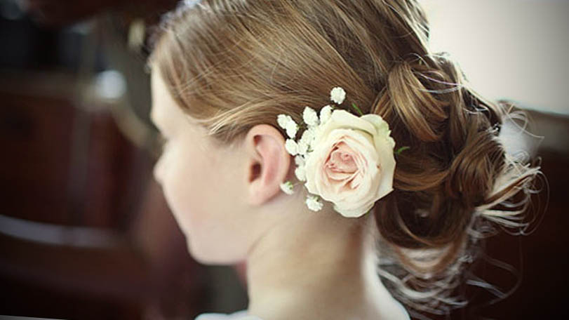 Close no penteado de coque baixo de uma daminha, com um arranjo de pequenas rosas brancas preso acima do coque.