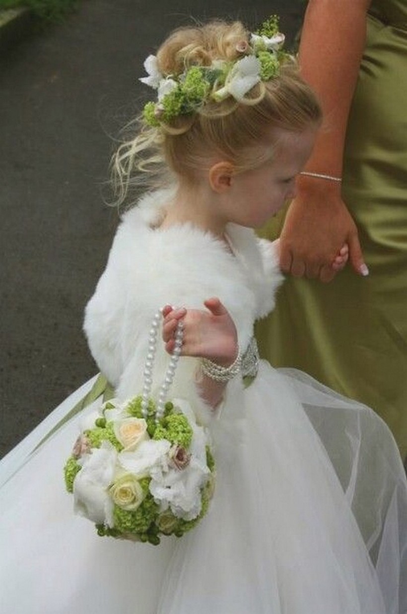 Daminha de costas com coque alto, coroa de flores brancas e folhagens, e um vestido com uma faixa verde na cintura.