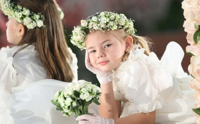 Daminha sorrindo, usando um vestido branco com luvas e uma coroa de flores brancas e folhagens no cabelo.