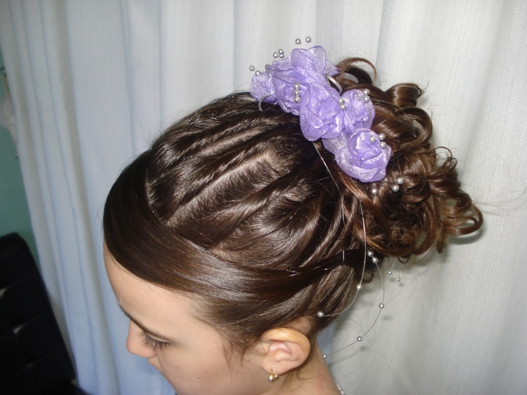 Daminha com penteado de coque cacheado alto, franja lisa e um arranjo de flores lilás preso ao lado do coque.